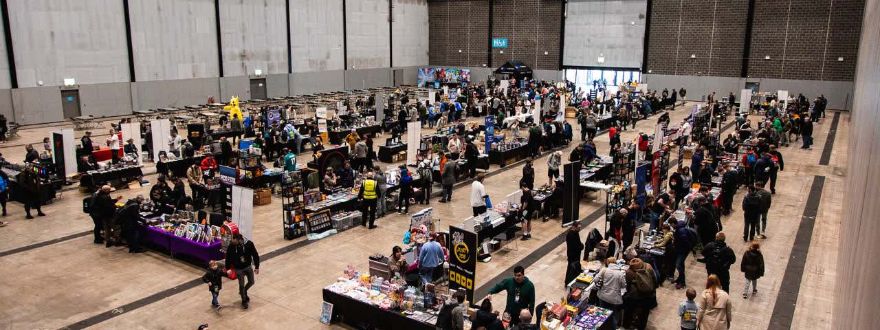 A high view over the hall floor, long rows of trading tables and people moving between them.