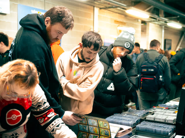 Collectors crowded round a table, one in a Poke Ball jumper holding a stack of cards.