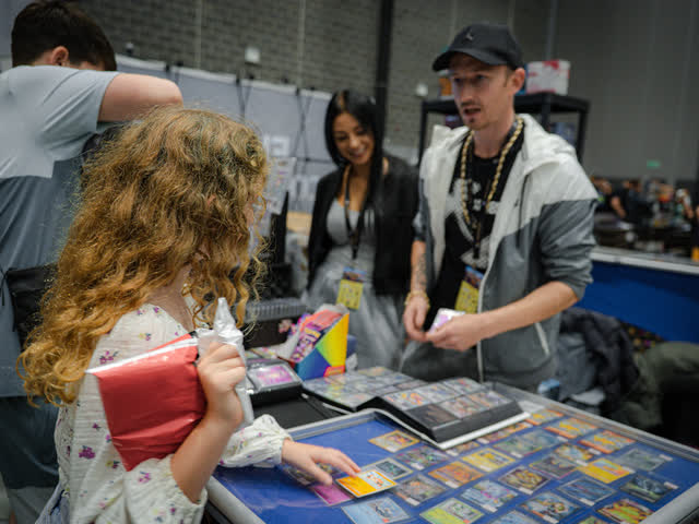 A girl holding a binder at a table while the trader behind it talks her through a card.