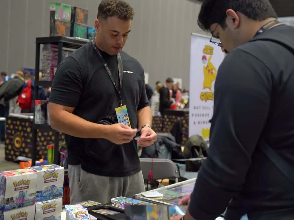 A vendor working his stand at a buy, sell and trade banner with the busy hall stretching away behind him.