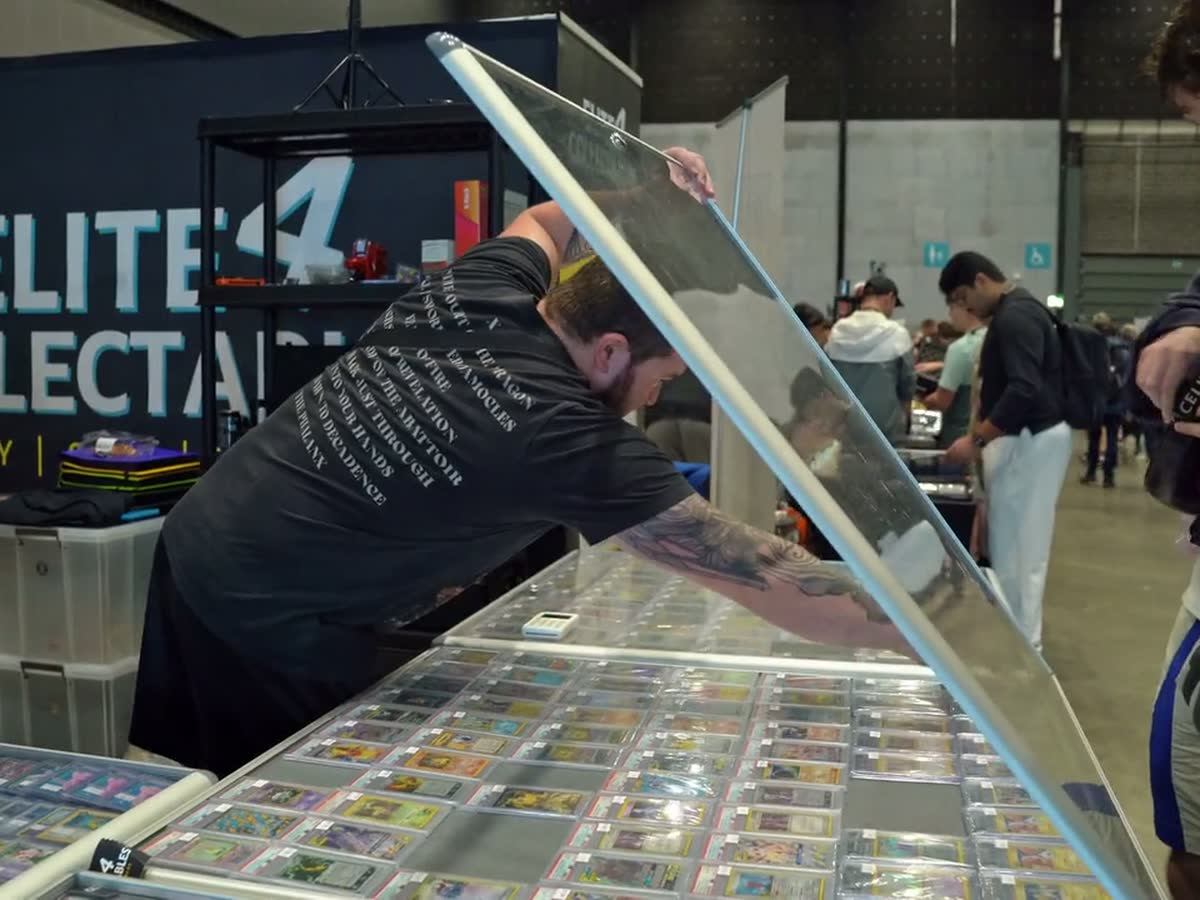 A trader lifting the lid of a glass case of cards at his stand, the hall busy behind him.